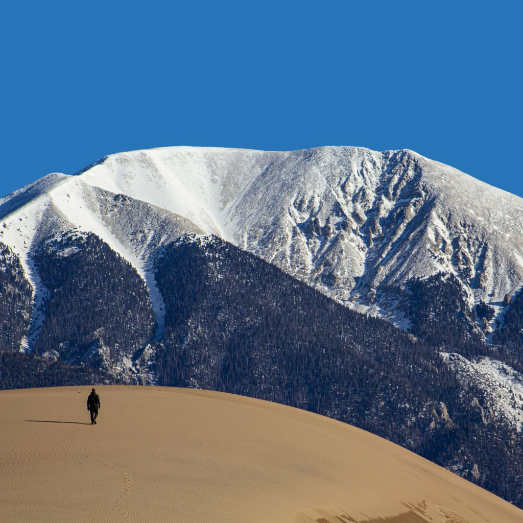 A lone hiker on a sand dune beneath a snowy mountain